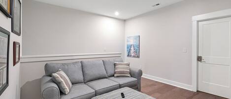 Una sala de estar con un sofá gris, un otoman tejido a rayas y una puerta a la derecha.