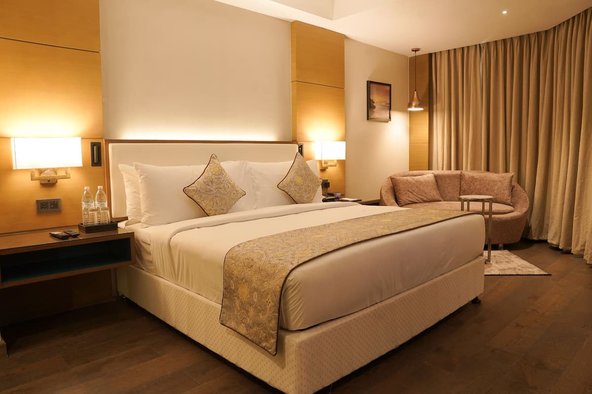 Ein Hotelzimmer mit einem großen Bett, Nachttischlampen, einem Nachttisch, einem Sessel und einem großen Fenster mit Vorhängen.