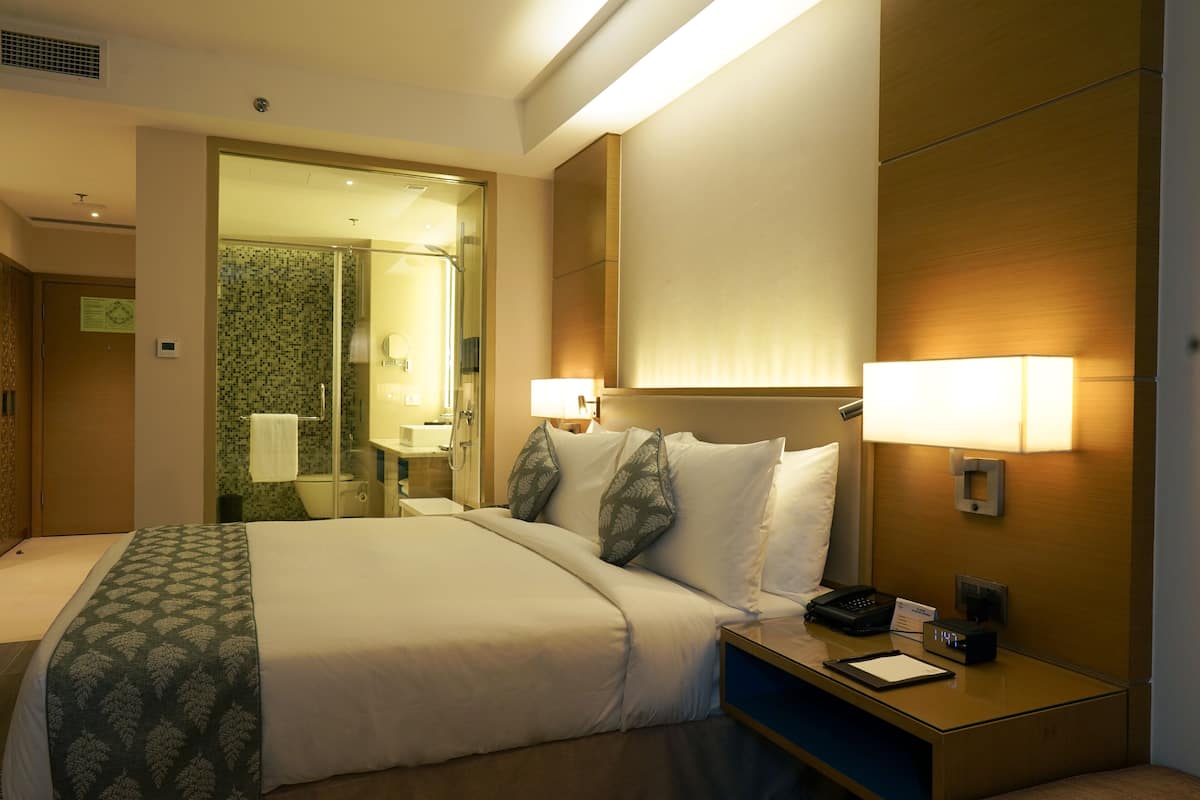 Ein Hotelzimmer mit einem großen Bett, Nachttischlampen, einem Nachttisch mit Telefon und Notizblock sowie einem durch eine offene Tür einsehbaren Badezimmer.