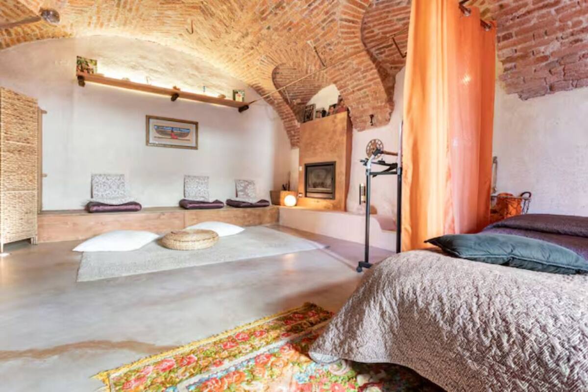 Une pièce chaleureuse avec un plafond en briques, un lit recouvert d’une courtepointe colorée, un petit coin salon avec des coussins et une étagère murale agrémentée d’objets décoratifs.
