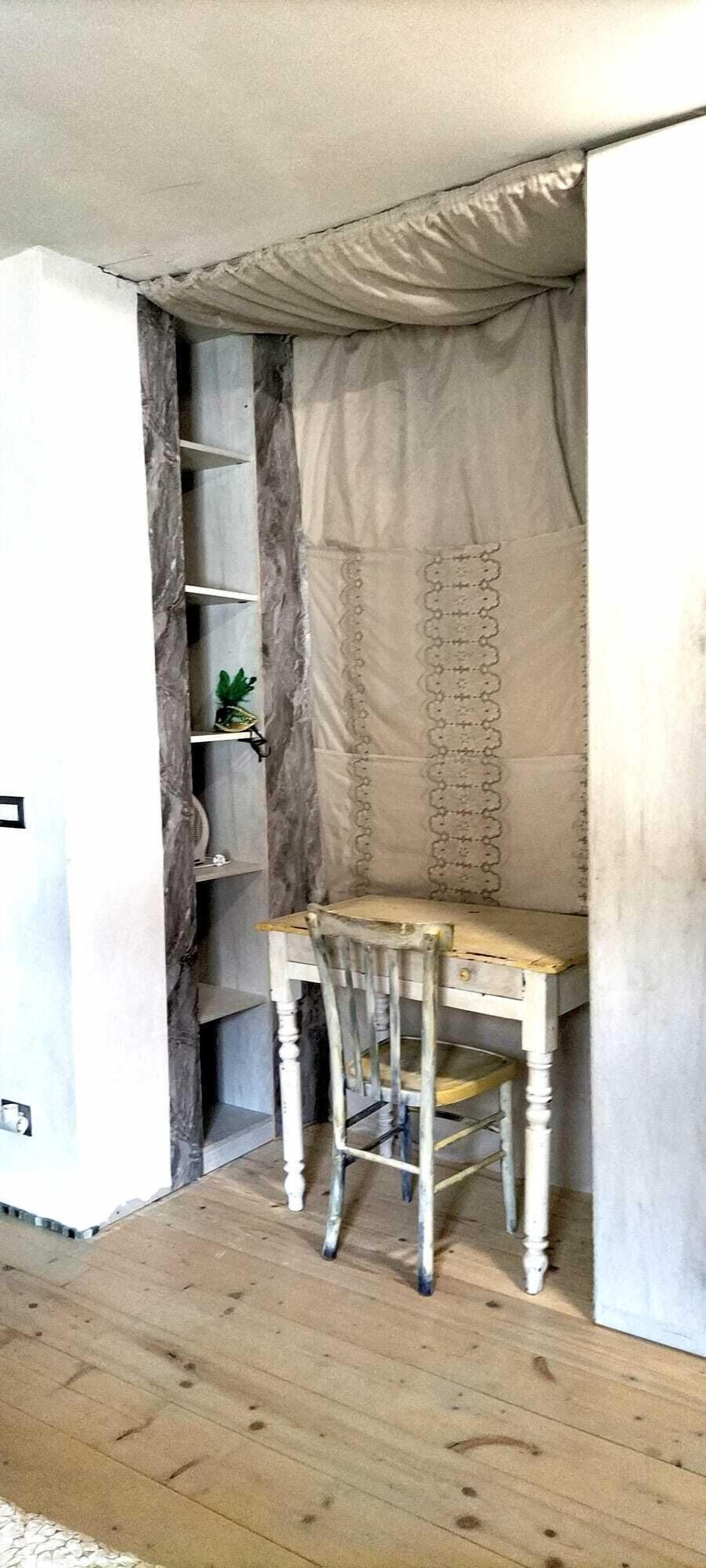 Une pièce comprenant un bureau en bois, une chaise, une étagère avec des objets décoratifs et un rideau.