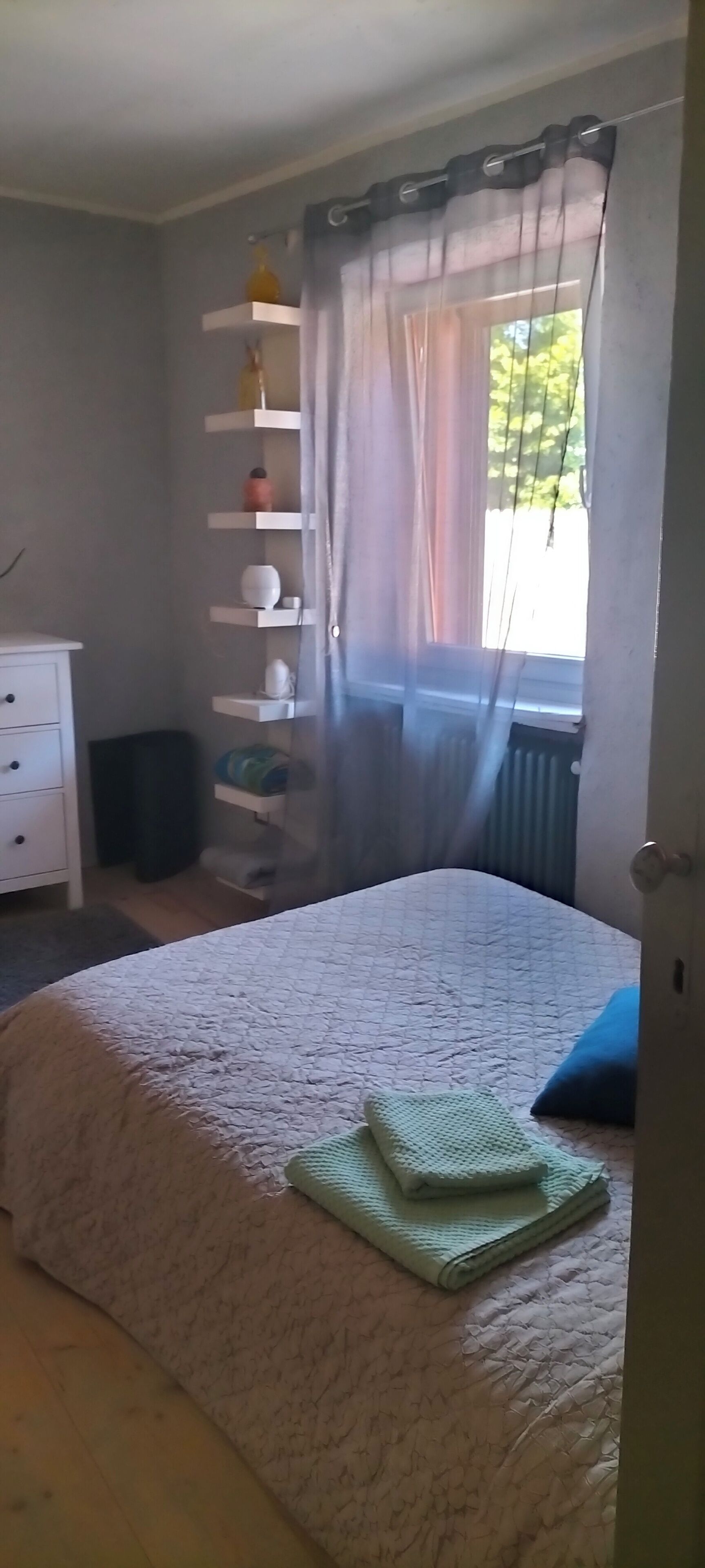 A bedroom with a bed, a bedside table, a dresser, a window with curtains, and a shelf with items.