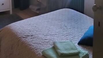 A bedroom with a bed, a bedside table, a dresser, a window with curtains, and a shelf with items.