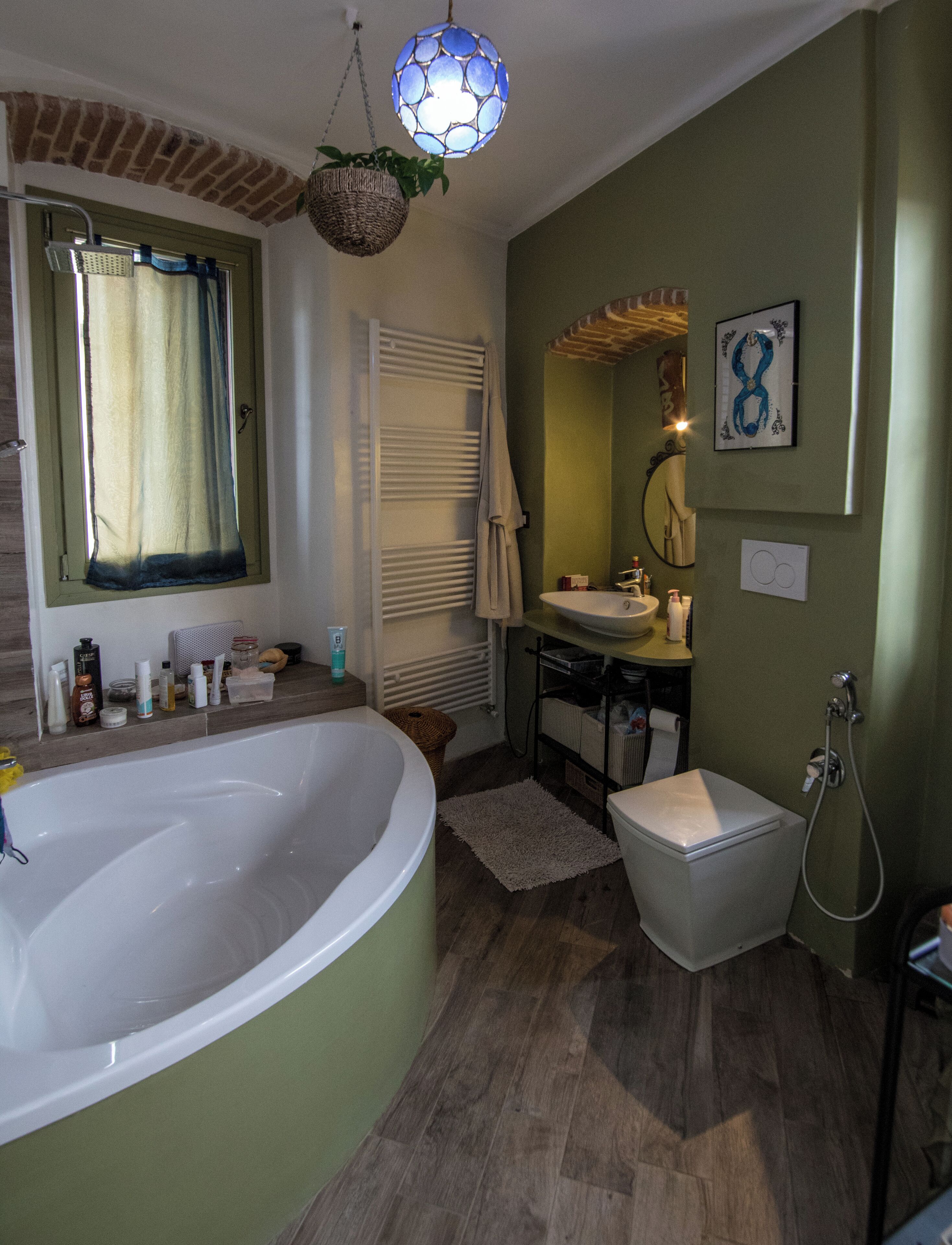 Un baño con bañera, lavamanos e inodoro. Cuenta con una ventana con cortinas, un perchero para toallas y una lámpara colgante.