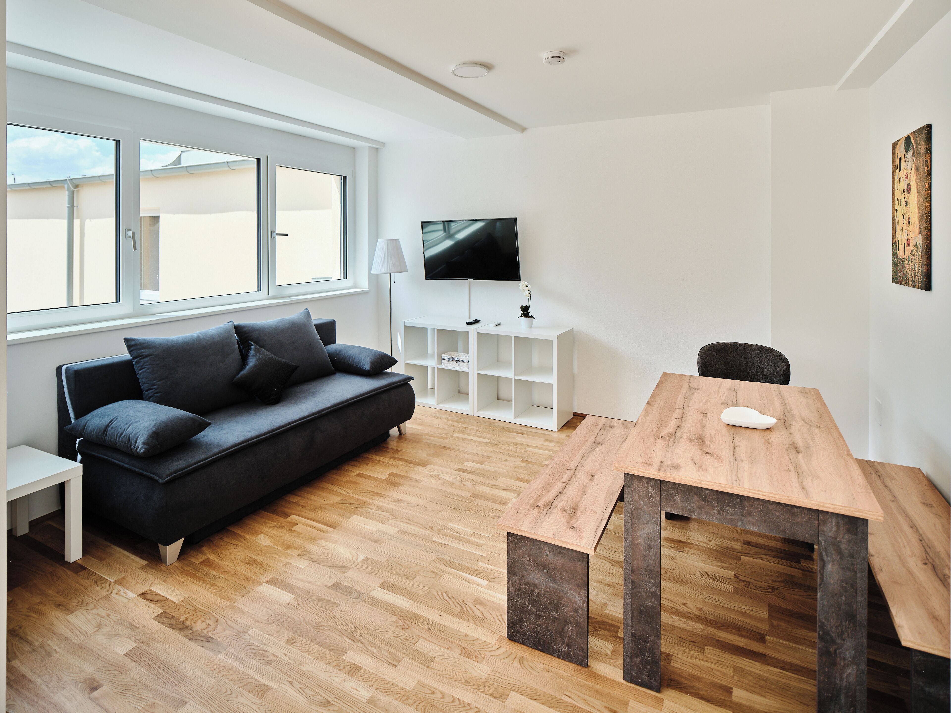A modern living room with a black sofa, a wooden dining table, and a TV on the wall.