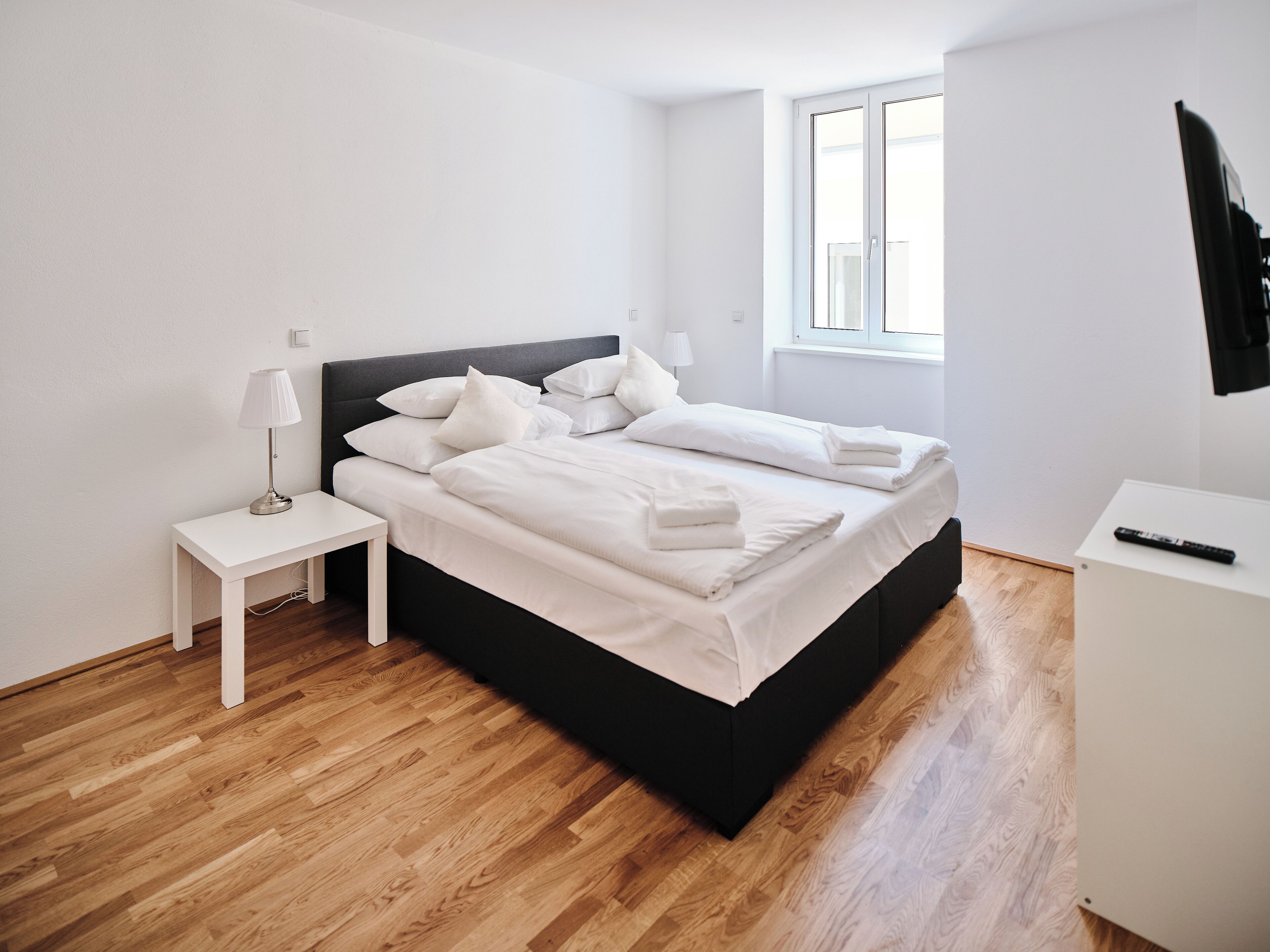 A modern bedroom with a black bed frame, white bedding, a bedside table with a lamp, and a small white cabinet with a remote control.