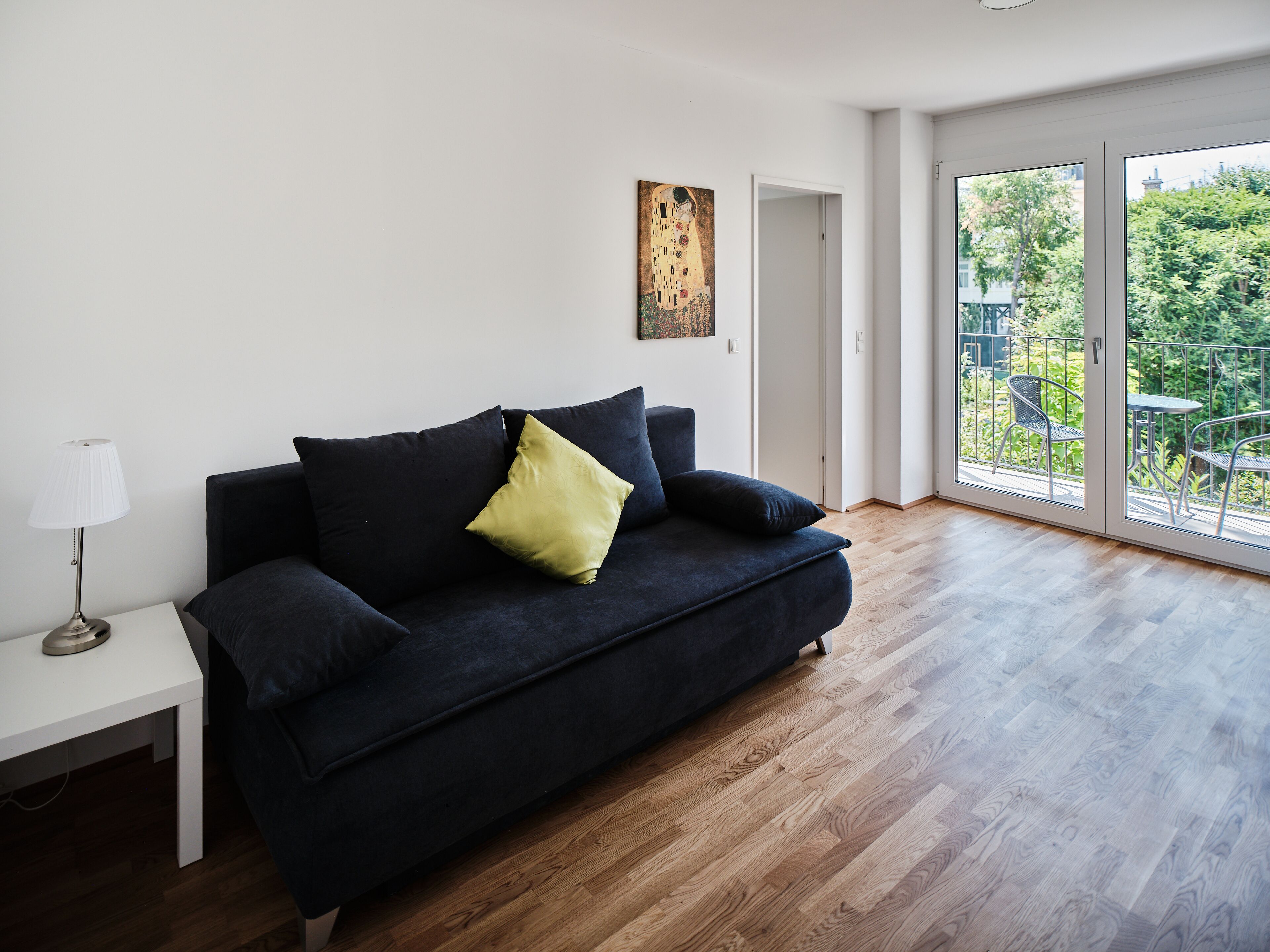 A modern living room with a dark blue sofa, a small white side table, and a floor lamp. There is a large window with a view of greenery outside.