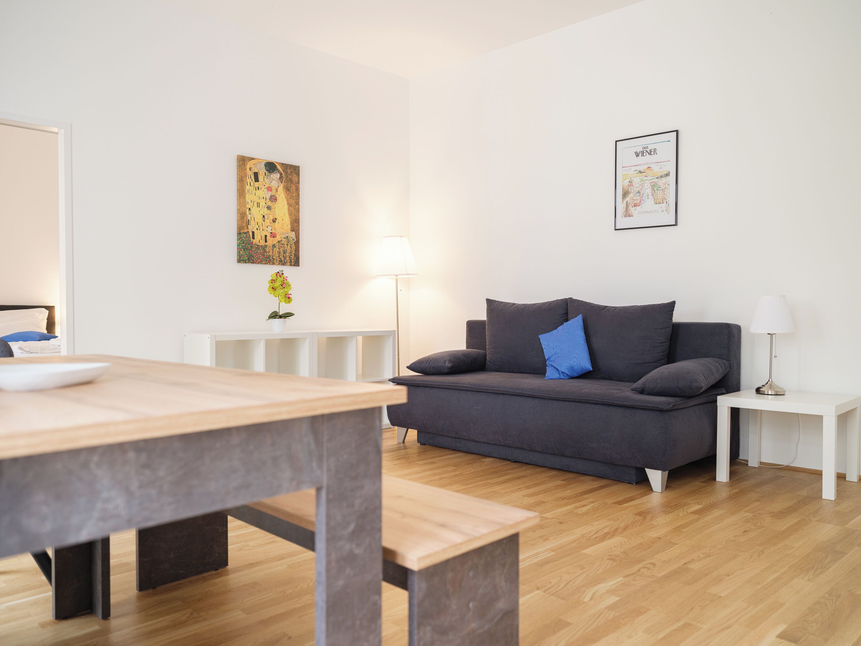 Un salon moderne avec un canapé gris, une table basse en bois, une table d’appoint blanche et un lampadaire. Un tableau encadré est accroché au mur et une petite plante en pot est posée sur la table d’appoint.