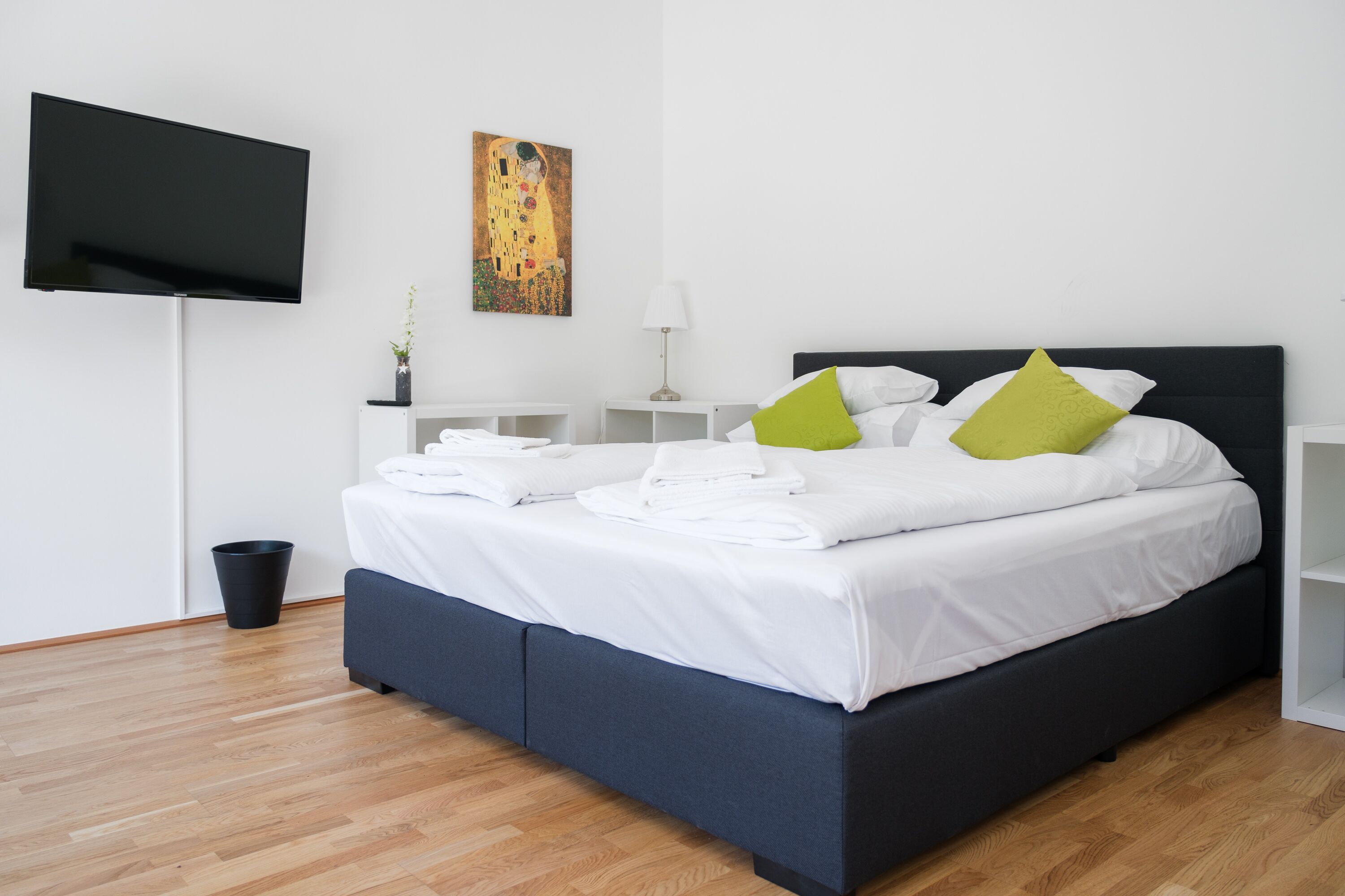 A modern hotel room with a large bed, a flat-screen TV, a painting, and a small bedside table with a lamp.