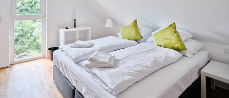 Une chambre à coucher moderne avec un grand lit, du linge de lit blanc et des coussins verts. On y trouve une petite table de chevet avec une lampe et un vase de fleurs.