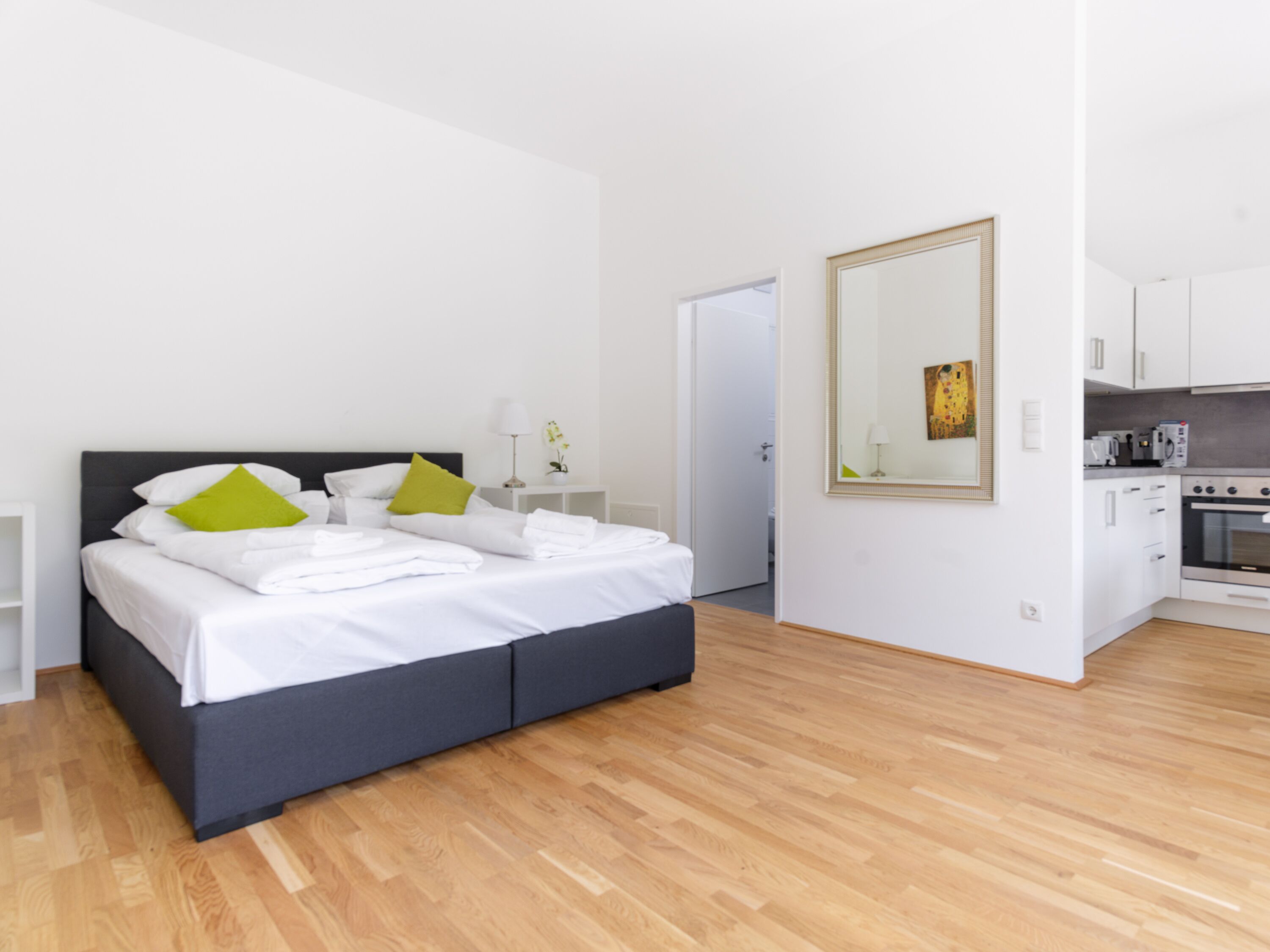 A modern hotel room with a large bed, a small bedside table, a mirror, a kitchen area, and a wooden floor.