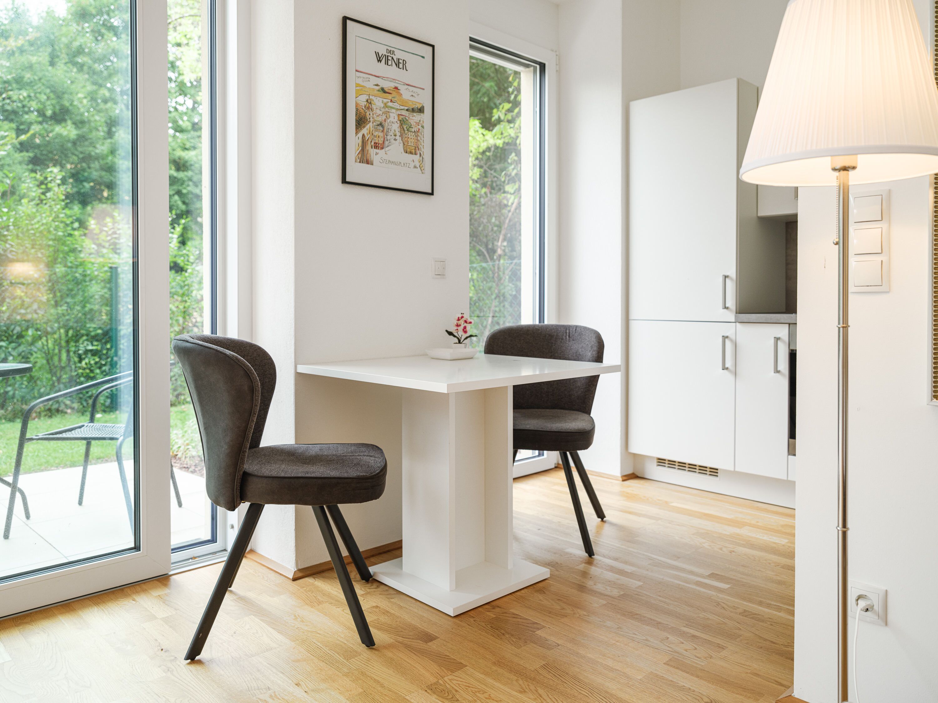 A modern kitchen with white cabinets, a white table, and two grey chairs. There is a window with a view of greenery outside.