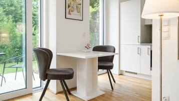 A modern kitchen with white cabinets, a white table, and two grey chairs. There is a window with a view of greenery outside.