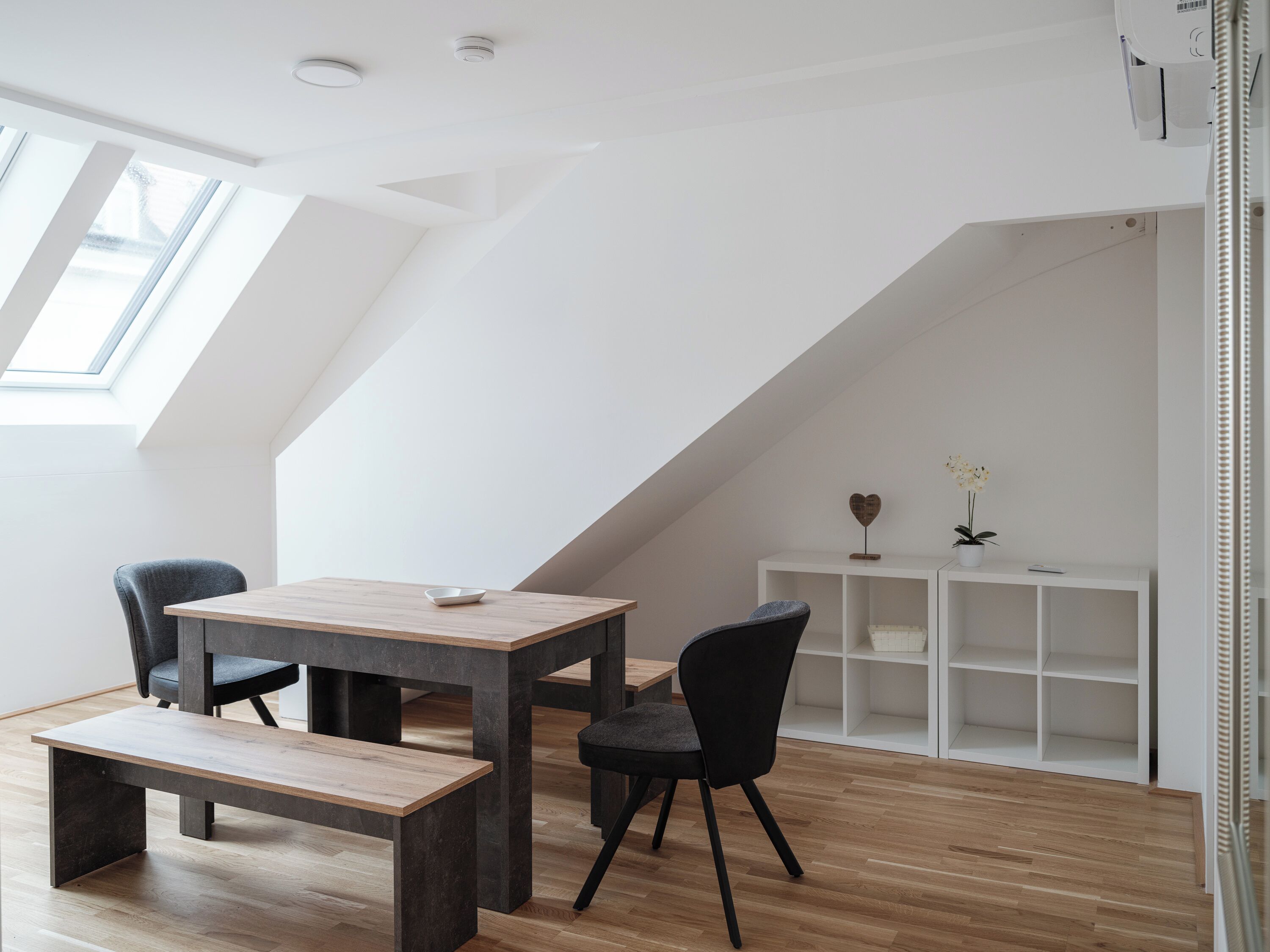 A modern loft bedroom with a sloped ceiling, skylight, wooden flooring, a central table with chairs, and a white shelving unit.