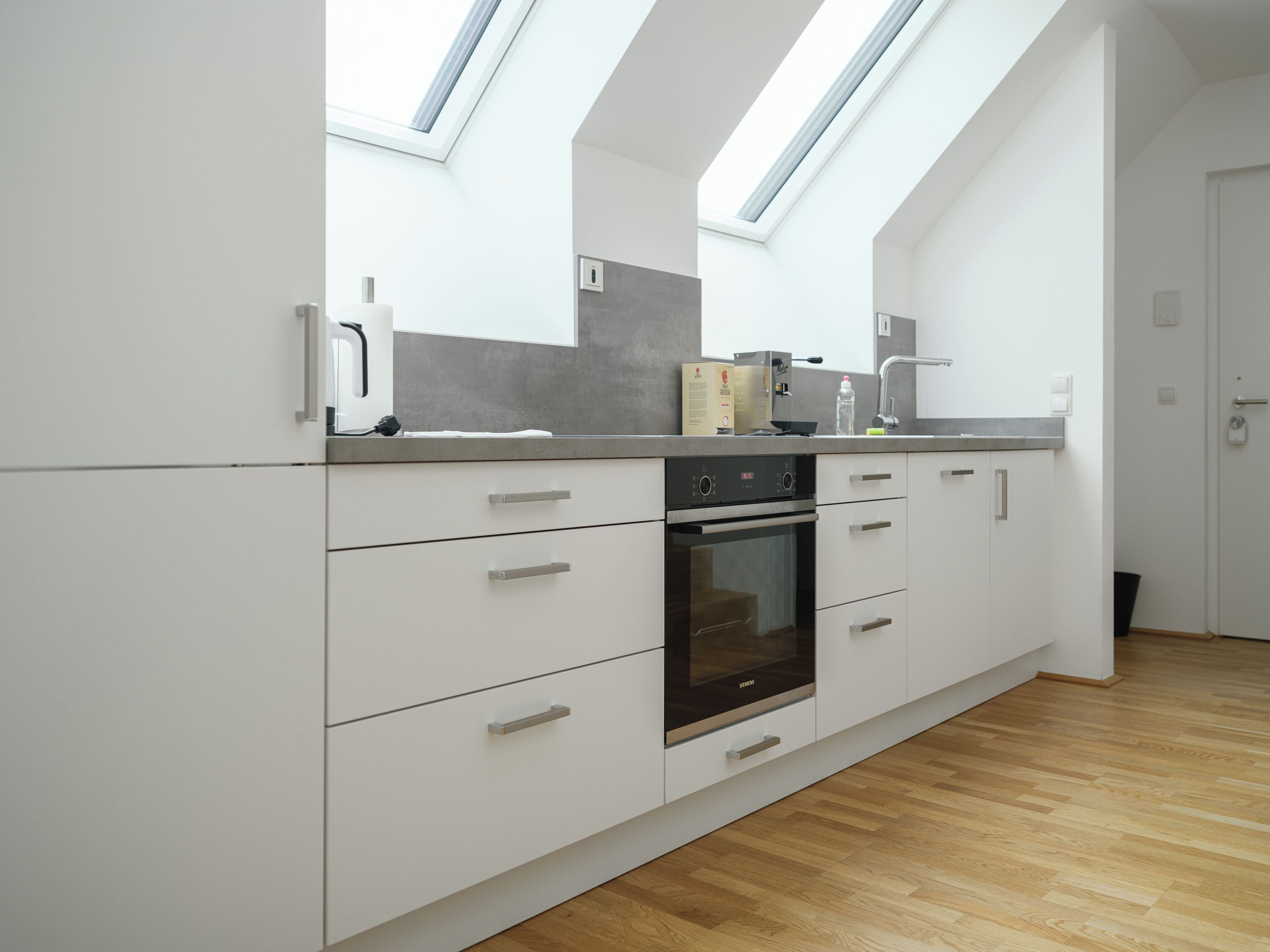 Une cuisine moderne dotée de fenêtres de toit, de meubles blancs, d’un four encastré et d’un plan de travail avec des appareils électroménagers.