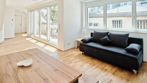A spacious living room with a wooden dining table, a dark sofa with cushions, and large windows offering a view of the outside. - My Schönbrunn Apartments (Vienna)