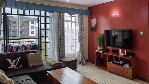 A living room with a sofa, coffee table, TV, and a balcony with clothes hanging. - Nice 3-bed Apartment in Nairobi (Nairobi)