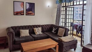 A living room with a dark brown sectional sofa, two framed artworks, a wooden coffee table, and a view of a balcony with a clothesline. - Nice 3-bed Apartment in Nairobi (Nairobi)