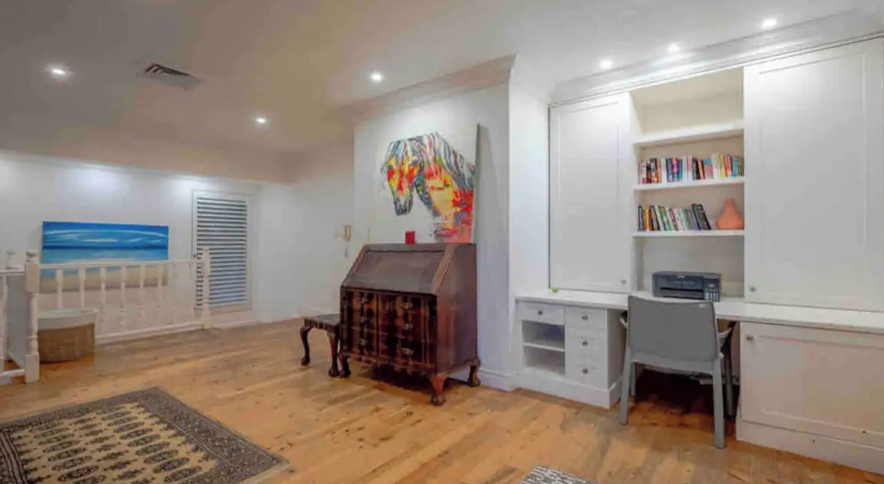 Una habitación amplia con suelo de madera, una estantería, un escritorio y un cuadro de colores en la pared.