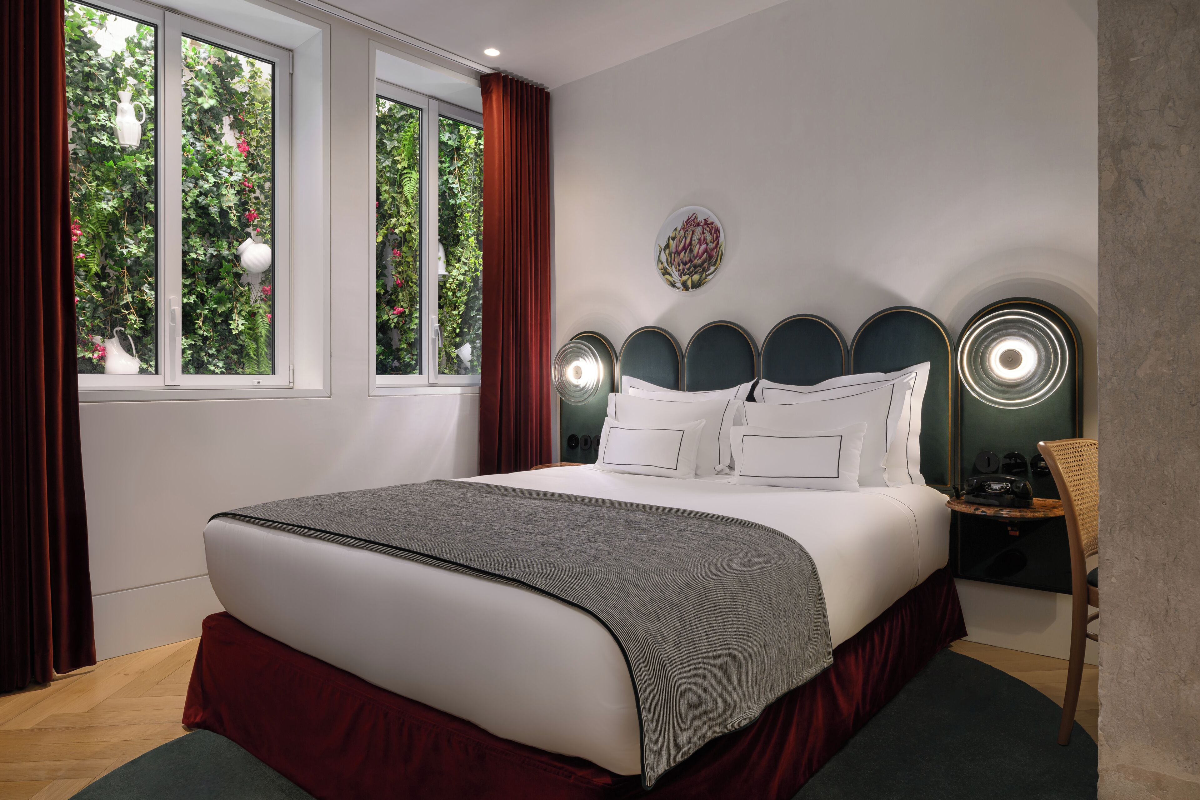 A bedroom with a bed, a nightstand, a chair, a window with curtains, and a wall-mounted light fixture.