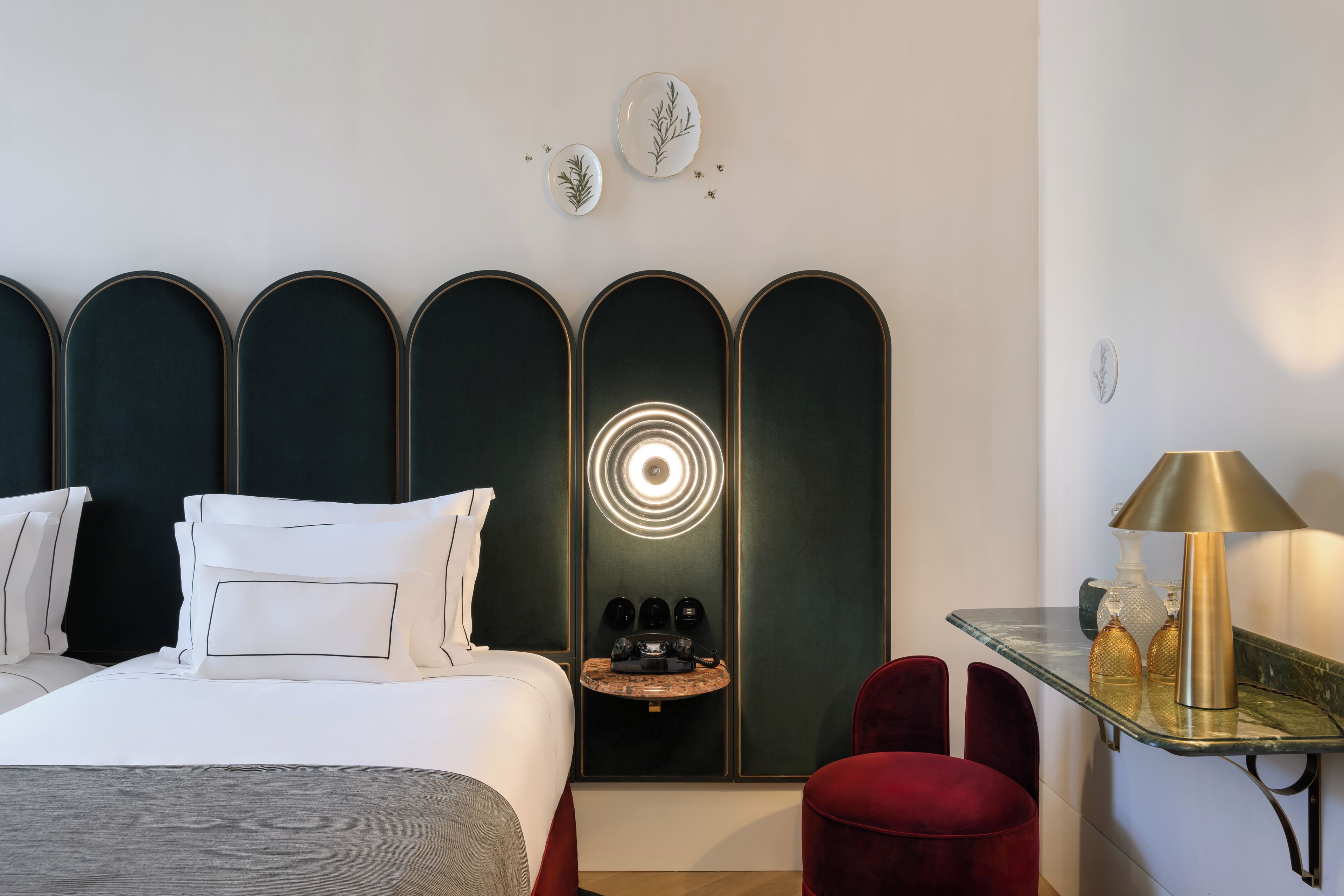 A hotel room with a bed, a desk with a lamp, a red chair, and a wall-mounted telephone.