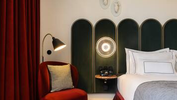 A hotel room with a red armchair, a bedside table with a lamp, a bed with white bedding, and a wall-mounted mirror.