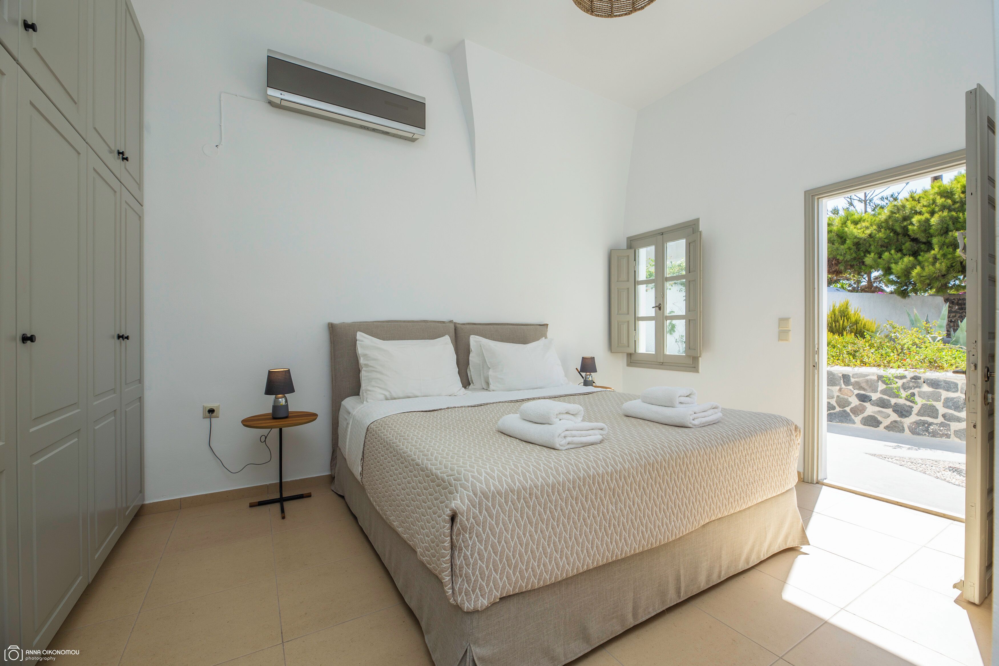 A bedroom with a bed, bedside table, lamp, air conditioning unit, and a view of the outdoors.