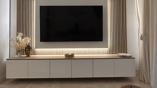 Una sala de estar moderna con un televisor de pantalla plana grande montado en una pared con persianas verticales, un mueble blanco debajo y una alfombra marrón grande en el suelo.
