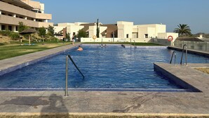 Una piscina con borde de azulejos, rodeada por un suelo de hormigón. Se observan palmeras y un edificio con techo plano al fondo.