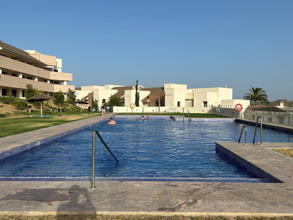 A swimming pool with a tiled edge, surrounded by a concrete deck. There are palm trees and a building with a flat roof in the background. - Apartamento Zen Garden. Valle del Este Golf (AN)
