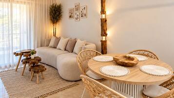A modern living room with a round wooden dining table, wicker chairs, a sofa, and a large window with sheer curtains.