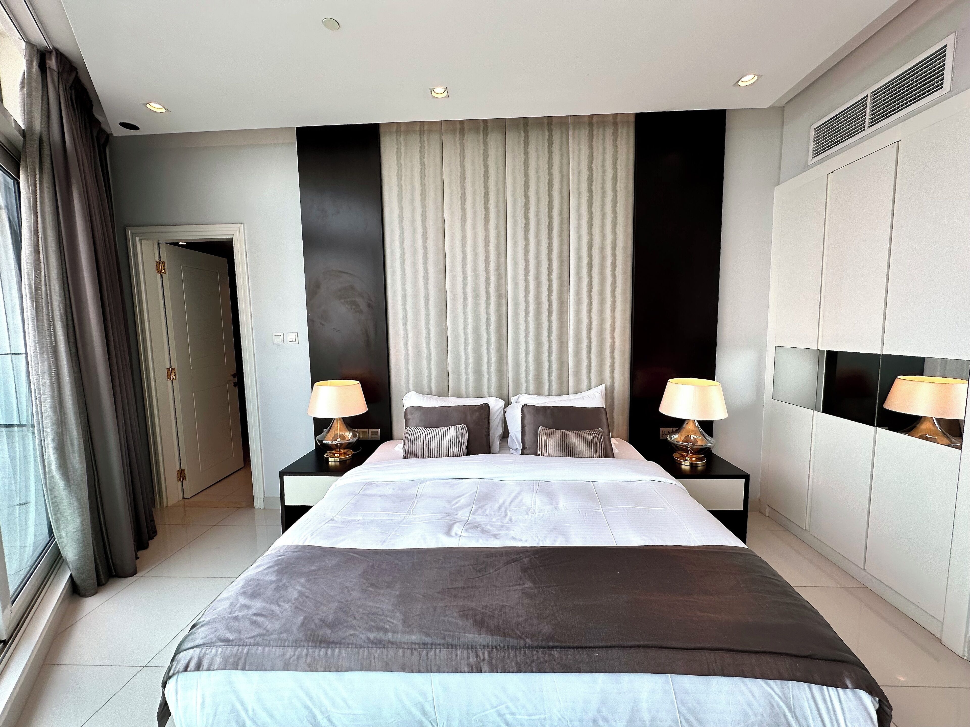 A modern hotel room with a large bed, two bedside tables with lamps, a window with curtains, and a door.