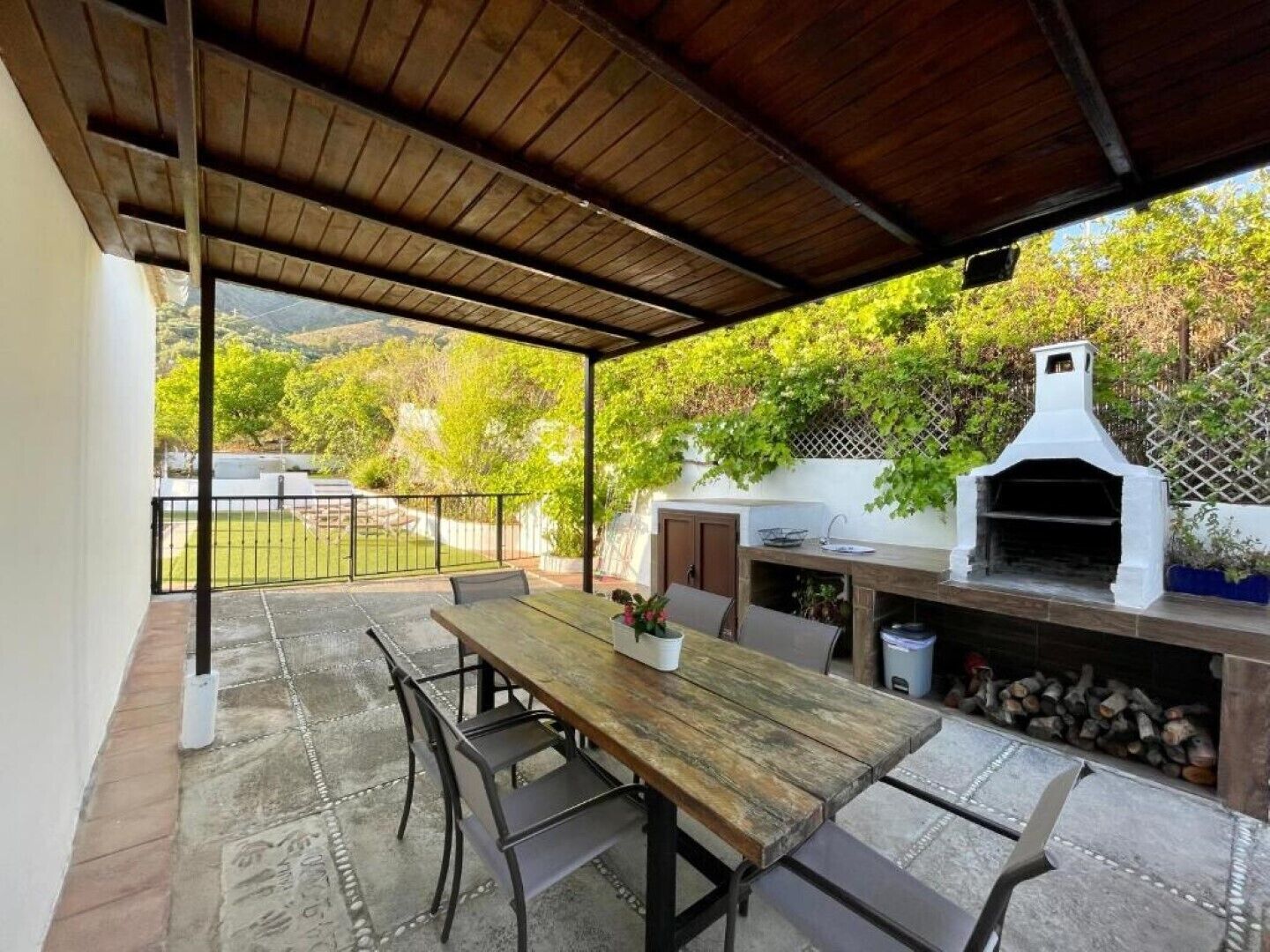 Een terras buiten met een houten tafel en stoelen, een barbecue en uitzicht op een tuin en heuvels.