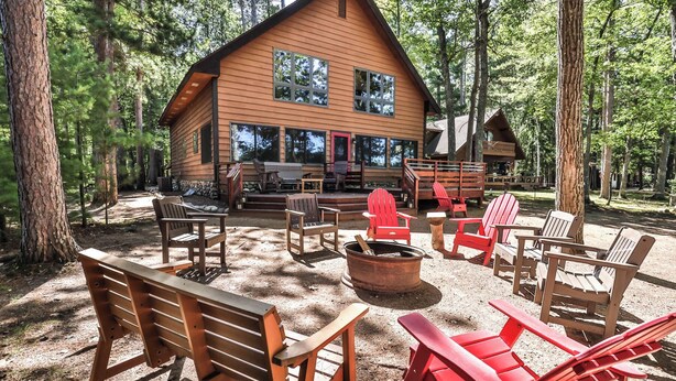 A cabin with a deck, surrounded by trees, and furnished with wooden chairs and a fire pit. - Stepaway Vacation Rentals - Shimmering Waters (St. Germain)