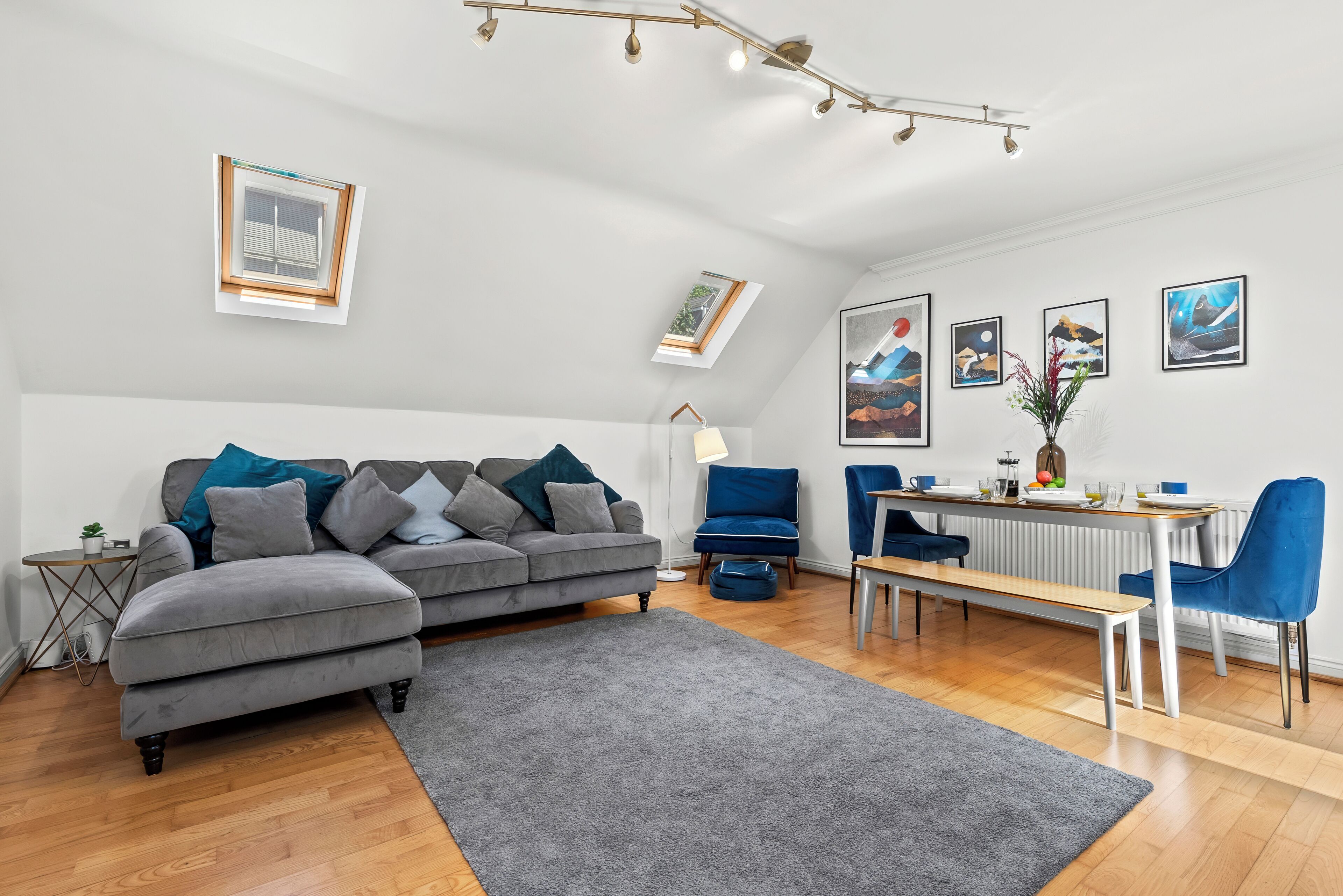 Un salon moderne avec un canapé gris, des fauteuils bleus, une table à manger et des œuvres d’art accrochées aux murs.