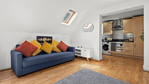 Ein modernes Wohnzimmer mit einem blauen Sofa, einem kleinen weißen Beistelltisch und einem grauen Teppich. Im Hintergrund ist eine Küche mit Holzausbau und einer Edelstahl-Dunstabzugshaube zu sehen.