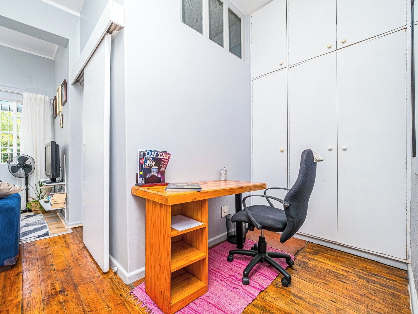A compact room with a wooden desk, a black office chair, a pink rug, and a white wardrobe.