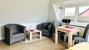 A modern room with a wooden floor, a desk with a computer, two black leather chairs, a small table with a red mat, and a bed with yellow bedding. - Haus Möwe App Austernfischer Westerland (Westerland)