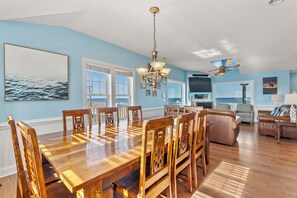 A dining area with a wooden table and chairs, a chandelier, and a view of the ocean through large windows.
