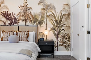 Ein Schlafzimmer mit einem Bett, einem Nachttisch, einer Wanduhr und einer Wand mit einem Wandbild von tropischer Vegetation.