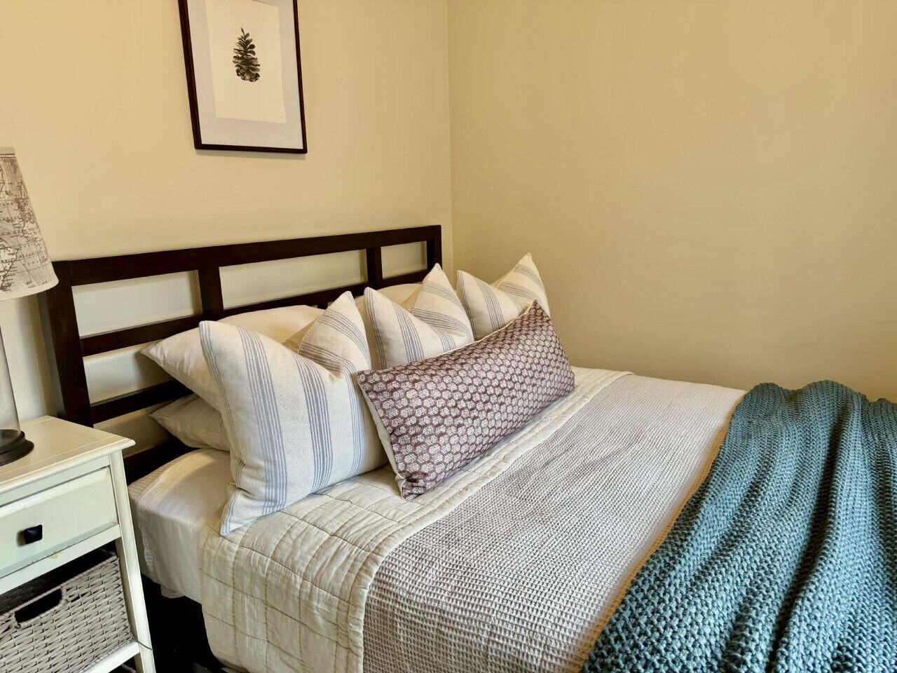 Un lit bien fait, avec une literie à rayures et un coussin à motifs, une table de chevet avec une lampe et un tableau encadré au mur.