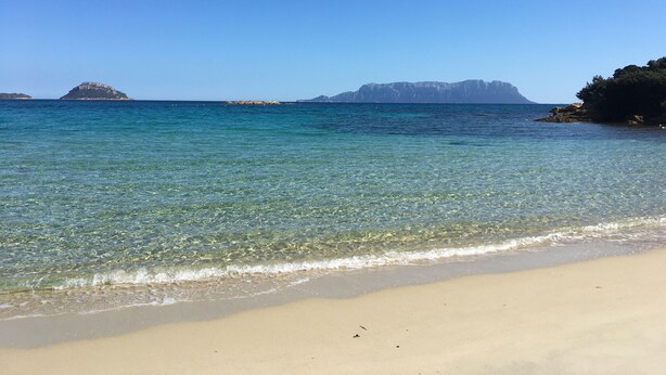 Una playa con agua cristalina, orilla de arena y unas islas rocosas a lo lejos.