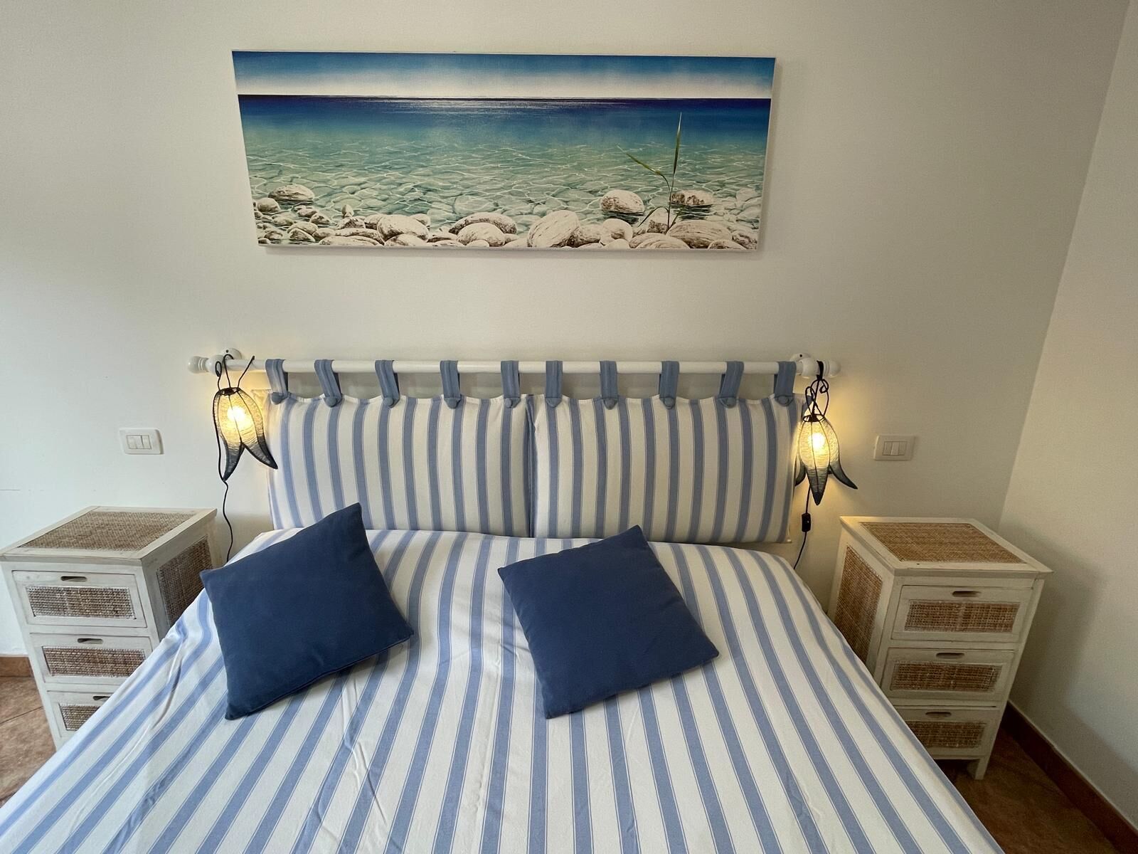 Camera da letto con coprip letto a righe, due cuscini, una testiera con tende e due abat-jour. Sul muro, un quadro raffigura una spiaggia.