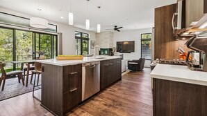 A modern kitchen with a large island, dark cabinetry, and a built-in oven. There is a dining area with chairs and a table, and a large window providing a view of the outdoors. - River West 814-Riverfront Silverthorne Condo, Fireplace, Hot Tub, Private Patio (Silverthorne)