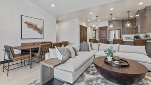 A modern living room with a sectional sofa, a dining area with a wooden table and chairs, and a kitchen with stainless steel appliances.