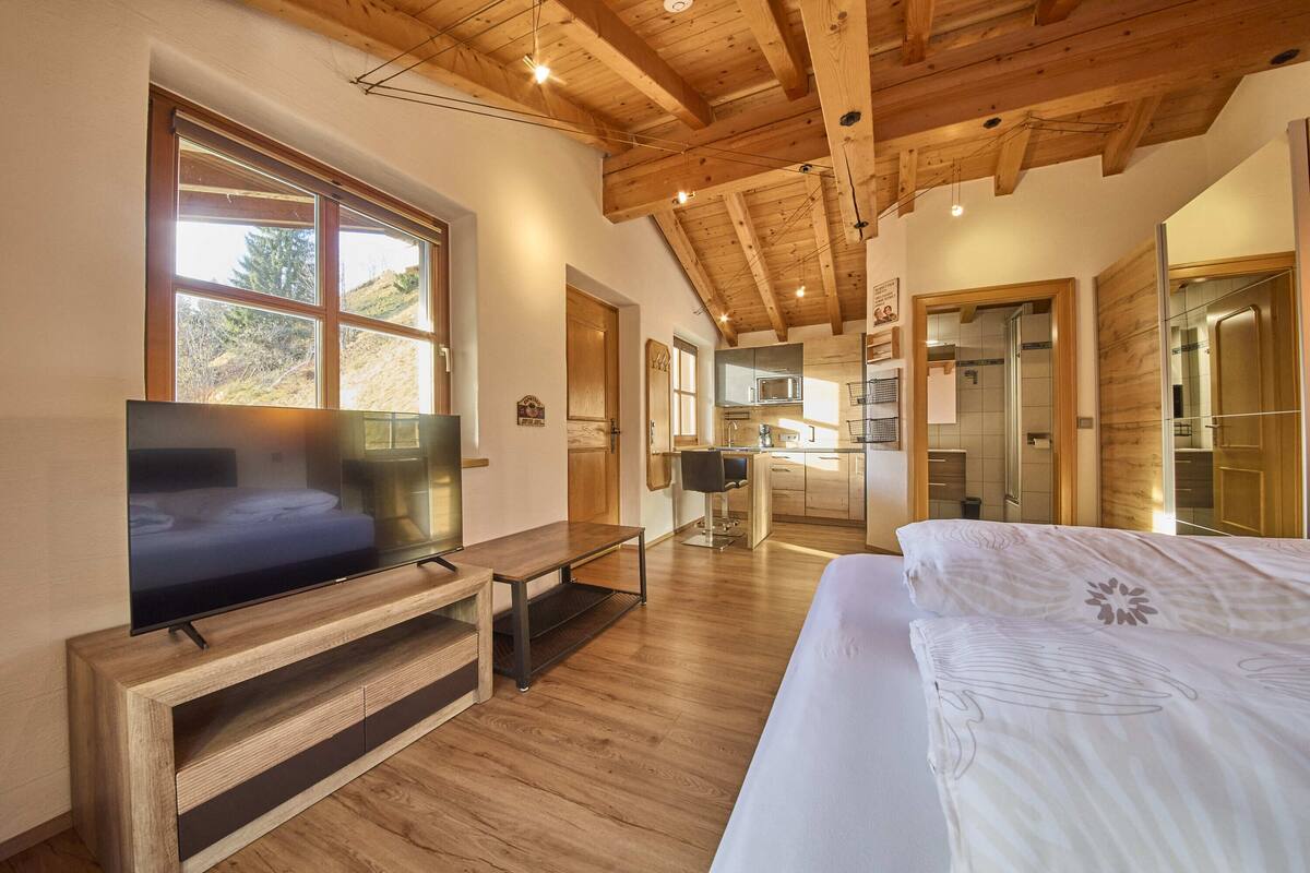 A modern hotel room with a wooden ceiling, a large bed, a TV, and a kitchen area visible through an open door.