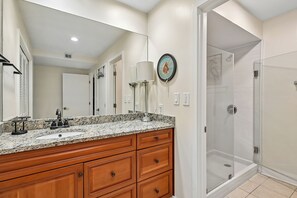 Un baño con encimera de granito, tocador de madera, un espejo grande, ducha con puertas de vidrio y una lámpara de pared.