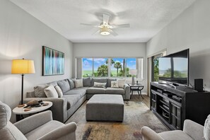 A modern living room with a sectional sofa, ottoman, TV, and ceiling fan.