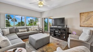 A spacious living room with a large gray sectional sofa, a gray ottoman, a flat-screen TV on a dark wooden stand, and a ceiling fan. - Oceanfront Villa w/ Private 2nd Floor Balcony (Hilton Head Island)