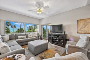 A spacious living room with a large gray sectional sofa, a gray ottoman, a flat-screen TV on a dark wooden stand, and a ceiling fan.